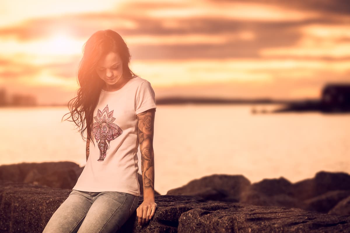 Kvinne i T-skjorte med elefanttrykk sitter på en stein ved havet i solnedgang